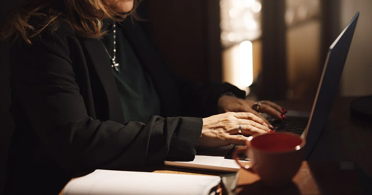A woman is focused on her laptop while typing in her home office. She has a cup of coffee and some notebooks on the desk. The light is dim, creating a cozy workspace.